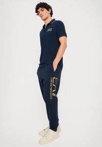 Marineblå polo shirt med guldaccent og logo, parret med matchende joggers med guldbranding, kompletteret med lyse sneakers.