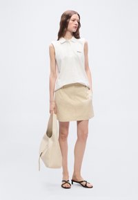 Γυναίκα με λευκή μαν sleeveless πόλο μπλούζα, μπεζ φούστα, μαύρα σανδάλια με τακούνι, κρατώντας μεγάλη μπεζ τσάντα tote, στέκεται μπροστά σε απλό φόντο.