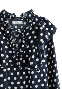 Marineblå bluse med hvide prikker, med rynket halsudskæring og ærmer, lavet af letvægtsstof. Etiketten er synlig i midten.