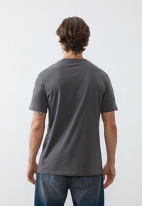Tricou gri cu mânecă scurtă, guler rotund, textură moale și croială lejeră, purtat cu blugi neavi. Vedere din spate care arată cusăturile umerilor.