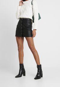 Mini-jupe en cuir noir avec fermeture à boutons à l'avant, associée à un pull en tricot torsadé crème. Le mannequin porte des bottines à talons noires et tient un sac vert.