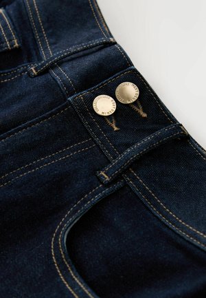 Jean en denim bleu foncé avec des boutons dorés, une couture dorée contrastante et un design classique à cinq poches. Texture lisse, coupe ajustée.