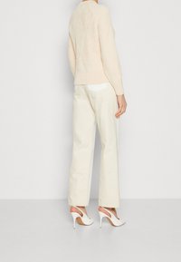 Kvinna i krämfärgad kabelstickad tröja, off-white raka jeans och vita slingback högklackade skor, stående mot en enkel ljus bakgrund.