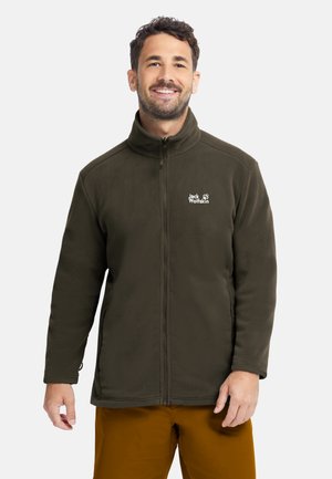 Uomo sorridente che indossa una giacca in pile verde scuro con zip e pantaloni marroni su uno sfondo chiaro e semplice.