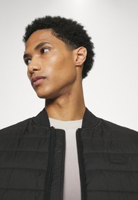 Giubbotto bomber nero con una texture trapuntata e colletto a costine, indossato sopra una camicia di colore chiaro. Zipper prominente e dettagli del logo ben visibili.
