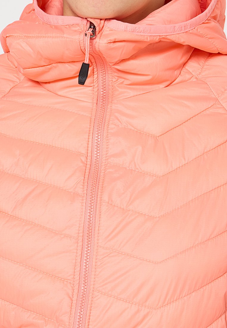 Chaqueta ligera, con capucha y acolchada en color rosa pastel. Presenta un cierre frontal con cremallera y un patrón en forma de chevrón en la tela. Textura suave.