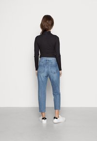 Žena otočená zády, oblečená v černém topu s dlouhým rukávem, vysokých modrých džínách a bílých teniskách s černými akcenty na podpatcích, stojí před bílou zdí.