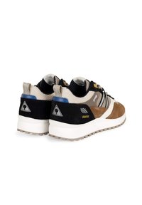 Braune und schwarze Sneaker aus Mesh- und Wildledermaterialien. Merkmale sind weiße Sohlen, gelbe Akzente und ein Dreieckslogo an der Ferse. Profilierte Außensohle.