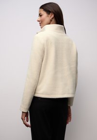 Kremowa kurtka wykonana z miękkiej, fakturowanej tkaniny, z wysokim kołnierzem i krótkim krojem. Cechuje ją minimalna ilość detali oraz czyste linie.