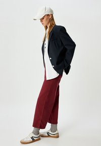 Zwarte blazer, witte overhemden, bordeaux brede broek, grijze sokken, witte sneakers met zwarte strepen en een witte pet.