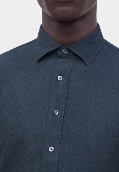 Gros plan sur le col et la poitrine d'une chemise bleu foncé à boutons, portée par une personne à la peau foncée, sur un fond clair uni.