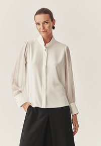 Weiße Bluse mit hohem Kragen, langen transparenten Ärmeln und knopfgefertigten Manschetten. Glattes Material mit leichtem Glanz, kombiniert mit schwarzen Hosen.