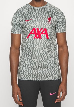 Homme portant un maillot à manches courtes gris à motifs du Liverpool FC avec le logo du sponsor AXA en rouge et le swoosh de Nike, associé à un pantalon noir avec un logo Nike rose.