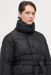 Čierna páperová bunda s vysokým golierom, zapínaním na gombíky, prešívanou textúrou a viazaným opaskom v páse. Hladký, lesklý materiál.