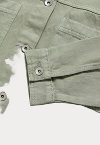 Giacca in denim verde chiaro con bottoni a pressione metallici, tasche frontali e una texture morbida. Il tessuto presenta un sottile motivo a trama.