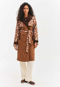 Brązowy pikowany płaszcz z beżowymi rękawami w cętki, szerokim kołnierzem i paskiem wiązanym w talii. W zestawie z kremowymi spodniami i brązowymi butami.