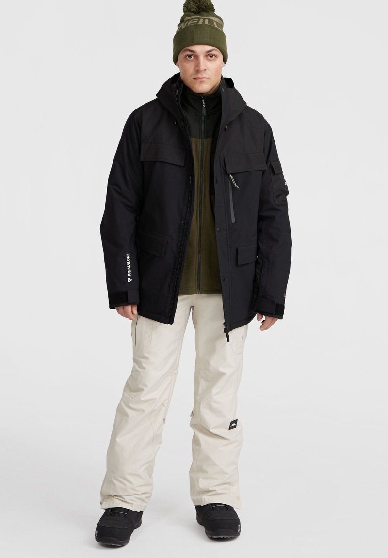 Czarna ocieplana kurtka z wieloma kieszeniami, w połączeniu z ciemnozieloną bluzą na zamek, kremowymi spodniami i czarnymi butami. Czapka beanie z logo na górze.