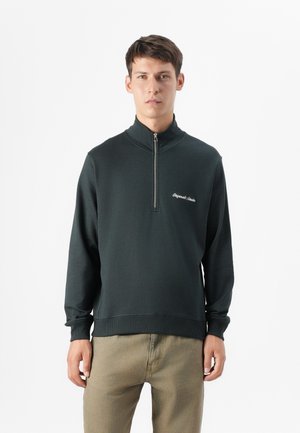 Model wears a dark green half-zip sweatshirt with "Original Style" embroidered on the chest, paired with light brown pants against a white background.