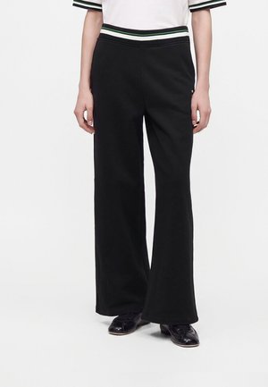 Personne portant un pantalon noir à jambes larges et des chaussures noires, avec une partie d'un haut blanc avec un bord rayé noir et vert.
