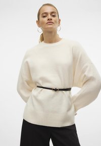 Cremfarbener Strickpullover mit Rundhalsausschnitt und geripptem Saum, tailliert mit einem dünnen schwarzen Gürtel mit goldfarbenem Akzent.
