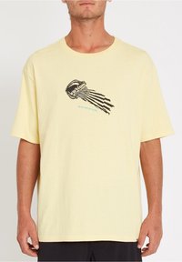 Mand iført en løs lys gul t-shirt med et sort og blåt vandmand grafik og teksten "Volcom" på brystet mod en hvid baggrund.