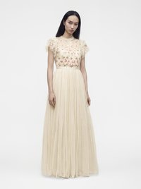 Femme aux longs cheveux noirs portant une robe beige longue jusqu'au sol avec une broderie florale sur le corsage et des manches courtes à volants.