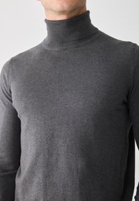 Hombre wearing un suéter de cuello alto ajustado gris oscuro, mostrando el cuello y la parte superior del torso contra un fondo claro y liso.