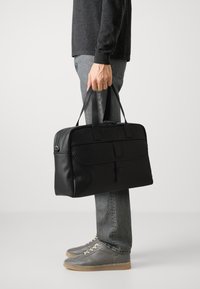 Bolsa rectangular negra con dos asas resistentes, textura de cuero suave, bolsillo con cremallera horizontal y diseño discreto.