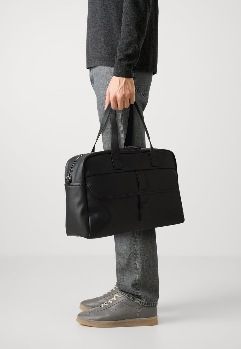 Bolsa rectangular negra con dos asas resistentes, textura de cuero suave, bolsillo con cremallera horizontal y diseño discreto.