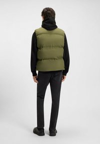 Chaleco verde acolchado sobre una sudadera negra con capucha, combinado con vaqueros oscuros y calzado negro. Presenta un diseño acolchado y un ajuste informal.