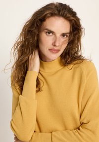 Frau mit lockigem braunem Haar, die einen senfgelben Strickpullover trägt, sanft ihren Hals berührt und direkt in die Kamera schaut.