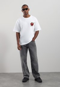 Weißes T-Shirt mit kurzen Ärmeln, das eine Grafik mit einem roten Herz und einem Schwert zeigt. Kombiniert mit locker sitzenden grauen Jeans und schwarzen Turnschuhen. Sonnenbrille getragen.