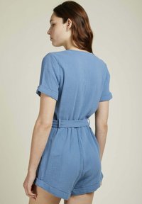 Combinaison en coton bleu avec des manches courtes, une taille ceinturée et des poignets retroussés. Le tissu texturé présente un motif de grille subtil.