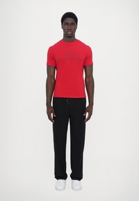 Raudona trumpa rankovė T-shirt marškiniai su juodu "ARMANI EXCHANGE" logotipu, derinami su juodomis kelnėmis ir baltomis sportbačiais. Paprastas, prigludęs dizainas.