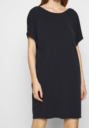 Femme portant une robe noire à manches courtes, ample, avec un large décolleté rond, debout devant un fond clair uni.