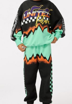 Persona con pantalones deportivos y sudadera a juego en negro y verde menta, con diseños en naranja de zigzag, tablero de ajedrez y texto "United Future".