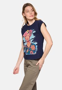 Mujer con top sin mangas azul marino con estampado colorido de flores y rostro de mujer, combinada con pantalones beige, de pie frente a un fondo blanco.