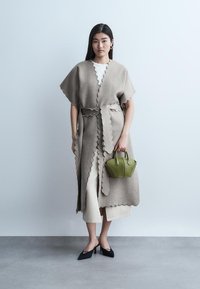 Žena u bež kaputu s nazubljenim rubom preko bijele odjeće, drži malu torbicu maslinasto zelene boje, stoji ispred jednobojne pozadine.