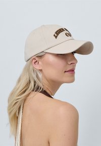 Beige sapka hímzett barna felirattal, strukturált dizájn, ívelt karima és állítható pánt hátul. Simább anyagtextúra.