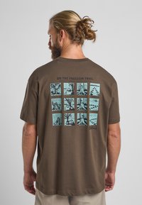 Bruine katoenen t-shirt met een grafiek op de achterkant met negen blauwe vierkanten die natuurtafereeltjes weergeven en de tekst "ON THE FREEDOM TRAIL."