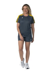 Top atletico grigio con dettagli gialli e maniche con motivo, abbinato a pantaloni sportivi grigi. Indossato con scarpe da ginnastica bianche.