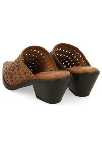 Mules à talons tressés marron avec un design ouvert, dotés d'une semelle intérieure en bois clair et d'un talon carré noir. Motif de tressage texturé visible.