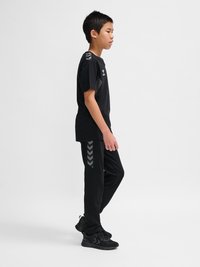 Garçon debout de profil portant une tenue de sport noire avec des motifs en chevrons sur les manches de la chemise et le pantalon, ainsi que des baskets noires.