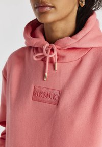 Primer plano de una persona que lleva una sudadera con capucha rosa, con un lazo de cordón y un logo bordado de "SIKSILK" en el pecho.