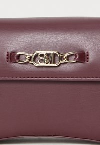 Bordeaux leren tas met een gladde textuur, voorzien van een gouden ketting en een logo-detail. Gestikte accenten voegen subtiele ontwerpelementen toe.