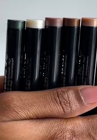 Viisi sylinterimäistä meikkitikkurasiaa eri sävyissä, mukaan lukien vihreä, valkoinen, ruskea ja pronssi, jotka pidetään käsissä, joiden iho on teksturoitu.