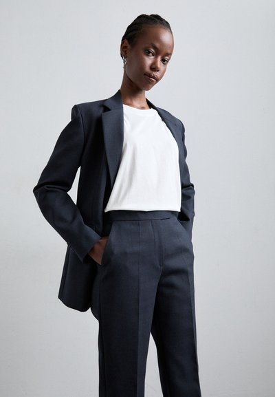 Blazer de marin cu umeri structurați, purtat peste un tricou alb. Pantaloni ajustați care se potrivesc cu blazerul, având linii clare și o textură netedă.