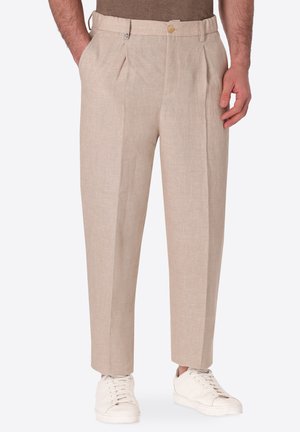 Uomo che indossa pantaloni beige plissettati con tasche laterali e sneakers bianche, in piedi davanti a uno sfondo semplice.