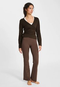 Brązowy top kopertowy z miękkiej tkaniny z długimi rękawami, zestawiony z brązowymi rozkloszowanymi legginsami. Oba elementy odzieży mają gładką fakturę.