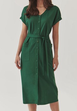 Vrouw die een groene jurk met korte mouwen en knopen draagt, met een strik in de taille en twee grote zakken aan de voorkant, staand tegen een effen achtergrond.